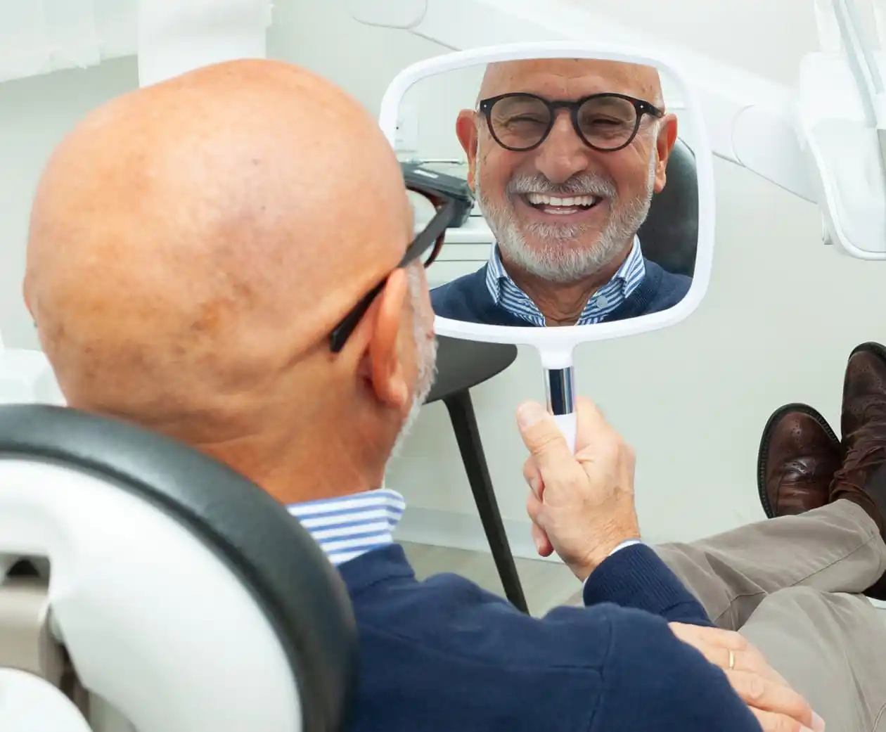 Older man sitting in an exam chair smiling into a mirror